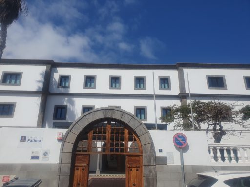 Centro de Nuestra Señora de Fátima Edificio blanco con puertas de madera y ventanas, bajo un cielo azul.